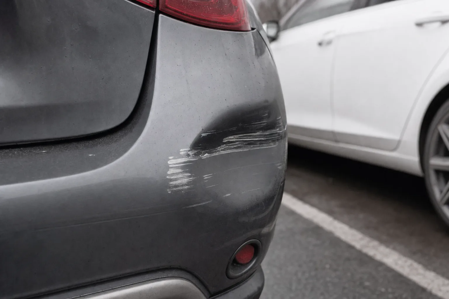Coin de pare-chocs gris fonce rayé avec un transfert de peinture blanche, une voiture blanche garée à côté.